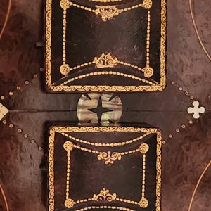 Puede incluir: Dos peque&ntilde;as cajas cuadradas de cuero marr&oacute;n con ribetes dorados y dise&ntilde;os ornamentados. Las cajas est&aacute;n sobre una superficie marr&oacute;n oscuro con un patr&oacute;n de peque&ntilde;os puntos blancos.