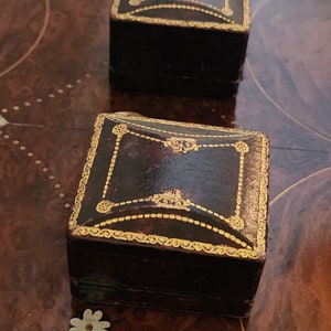 Puede incluir: Dos peque&ntilde;as cajas cuadradas de cuero negro con adornos dorados y detalles decorativos. Las cajas est&aacute;n sobre una superficie de madera.
