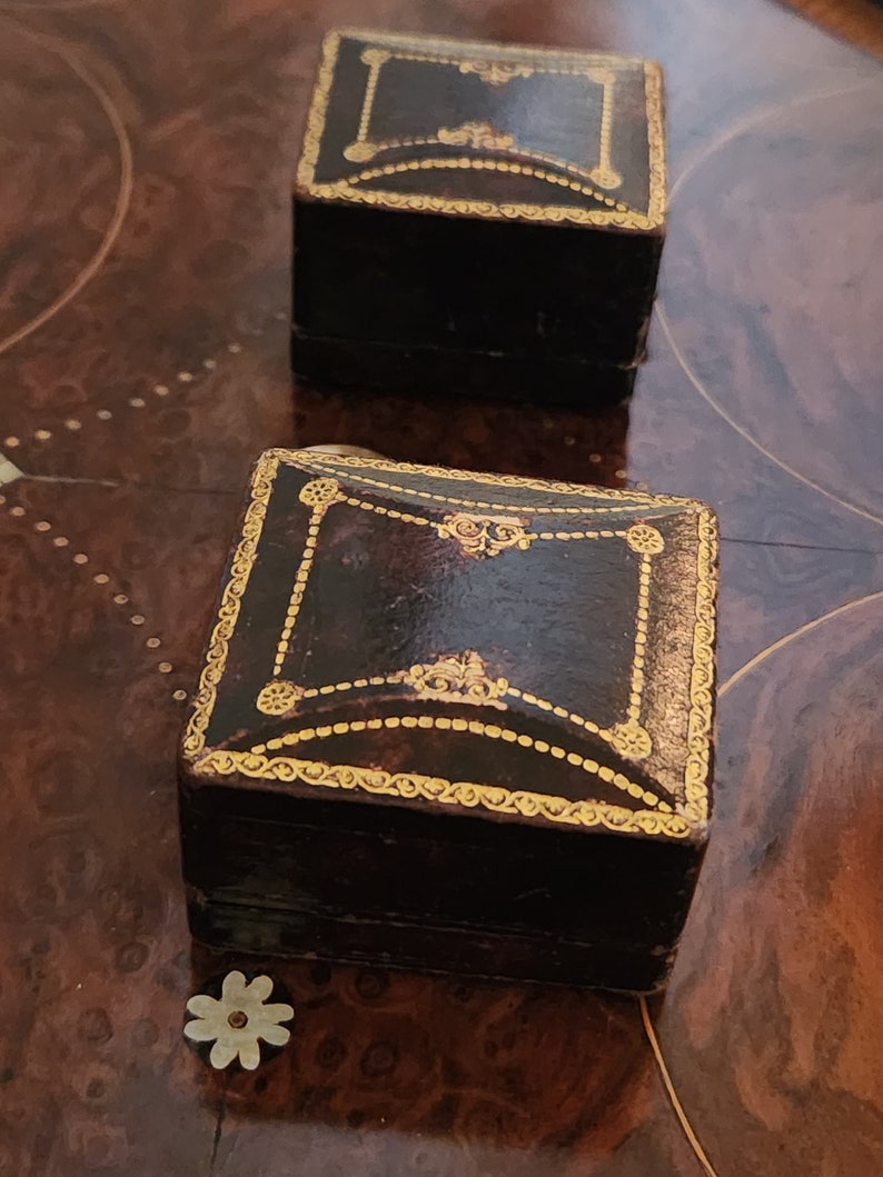 Puede incluir: Dos peque&ntilde;as cajas cuadradas de cuero negro con adornos dorados y detalles decorativos. Las cajas est&aacute;n sobre una superficie de madera.