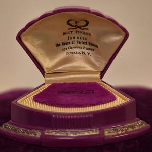 May include: A vintage purple jewellery box with a velvet exterior and a satin interior. The box is open, revealing a ring holder. The interior features the text "BERT FISCHER JEWELER The House of Perfect Diamonds 372 GENESEE STREET BUFFALO, N.Y."