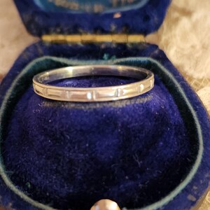 May include: A thin silver band with a simple, repeating pattern sits in a blue velvet ring box.