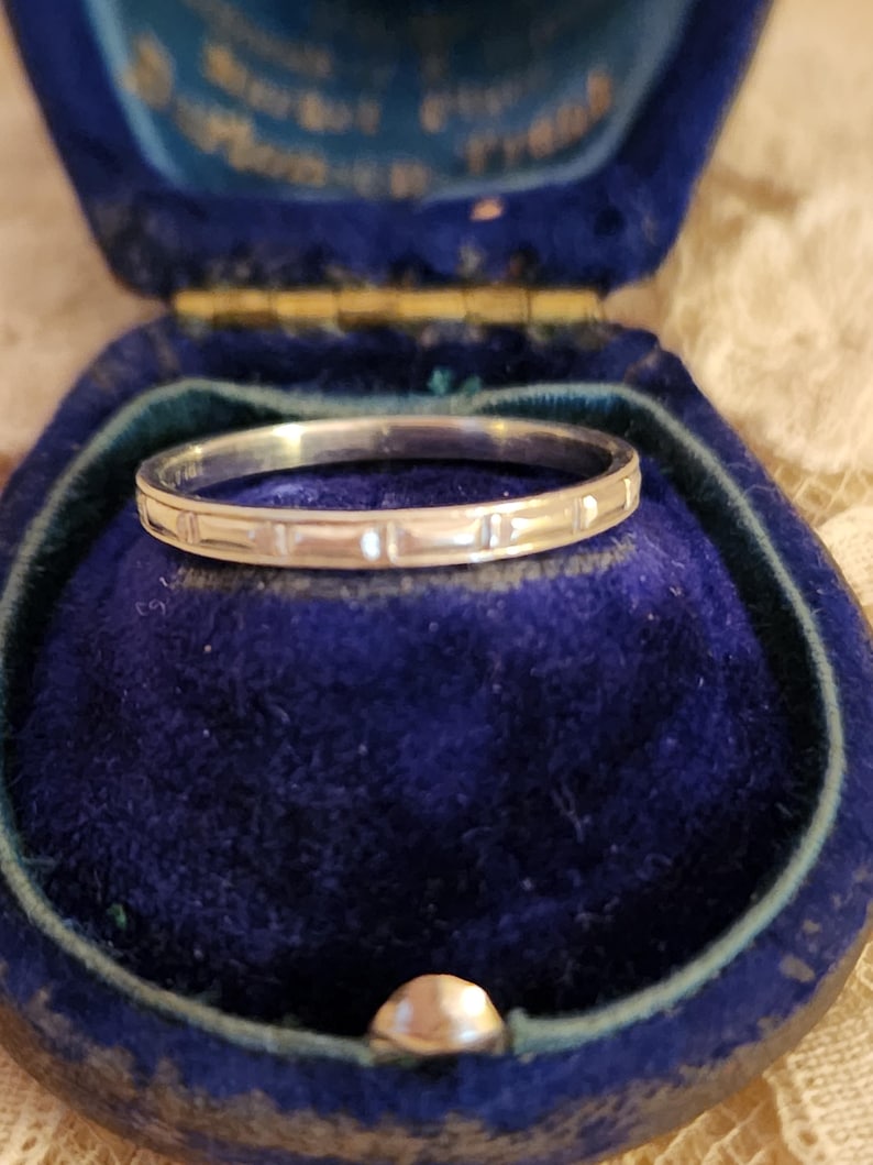 May include: A thin silver band with a simple, repeating pattern sits in a blue velvet ring box.
