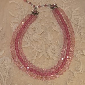 Könnte beinhalten: Eine mehrsträngige Halskette aus rosa Perlen. Die Halskette hat verschiedene Rosatöne, einen zarten Verschluss und dekorative Akzente. Die Halskette wird auf einer Spitze präsentiert.