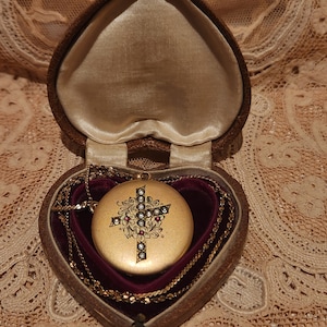 Pode incluir: Um medalhão de ouro com um design de cruz adornado com joias, dentro de uma caixa em forma de coração com um forro de veludo vermelho.