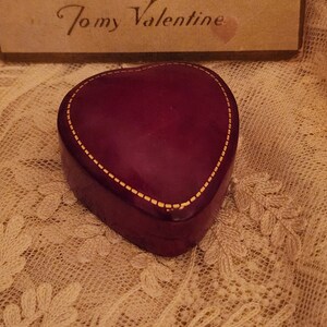 May include: A heart-shaped, burgundy-colored box with a gold trim. The box is sitting on a white lace surface. The text "To my Valentine" is visible on a card in the background.