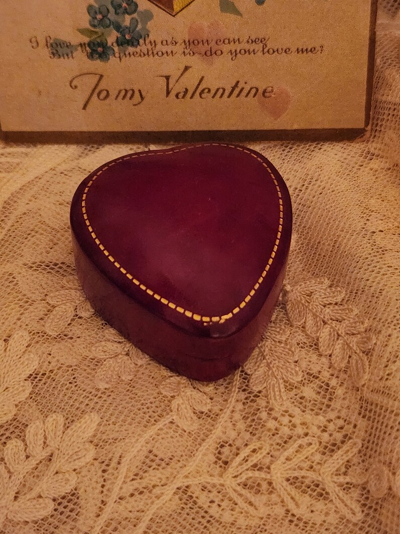 May include: A heart-shaped, burgundy-colored box with a gold trim. The box is sitting on a white lace surface. The text "To my Valentine" is visible on a card in the background.