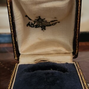 Puede incluir: Una peque&ntilde;a caja de anillo de color marr&oacute;n oscuro con un borde dorado y un interior de terciopelo azul. La caja est&aacute; abierta y el interior de terciopelo es visible.