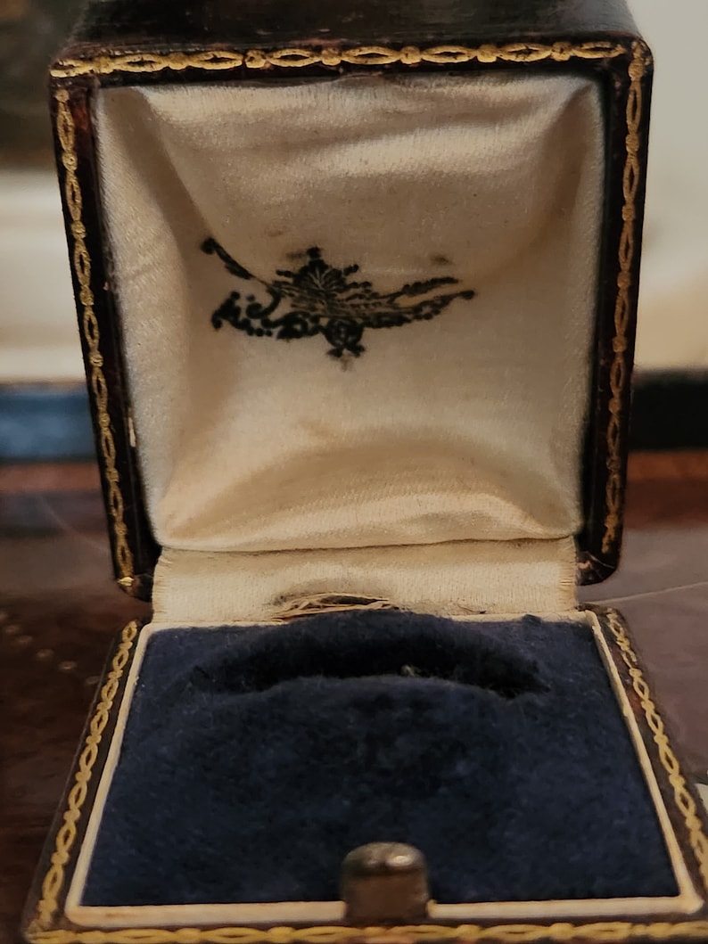 Puede incluir: Una peque&ntilde;a caja de anillo de color marr&oacute;n oscuro con un borde dorado y un interior de terciopelo azul. La caja est&aacute; abierta y el interior de terciopelo es visible.