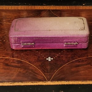May include: A small, purple velvet jewellery box with gold accents and a hinged lid. The box is closed and sitting on a wooden surface.