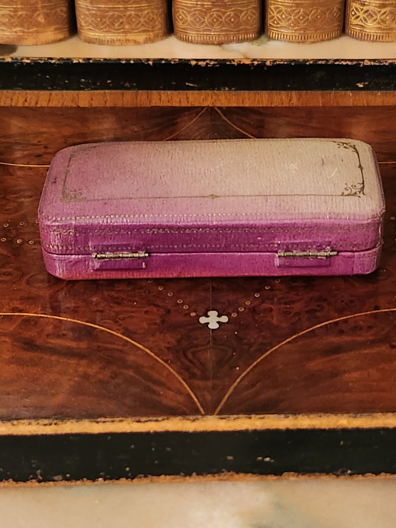 May include: A small, purple velvet jewellery box with gold accents and a hinged lid. The box is closed and sitting on a wooden surface.