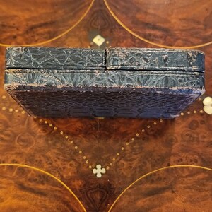 May include: A black leather box with a textured surface. The box is closed and sitting on a wooden surface.