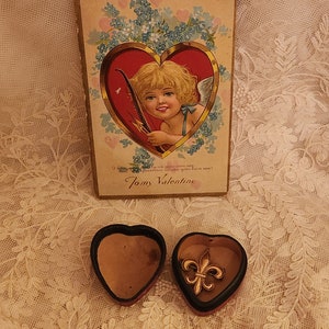 May include: A vintage Valentine's Day card with a cherub holding a bow and arrow, surrounded by flowers. The card is in a heart-shaped box with a gold fleur-de-lis charm inside.