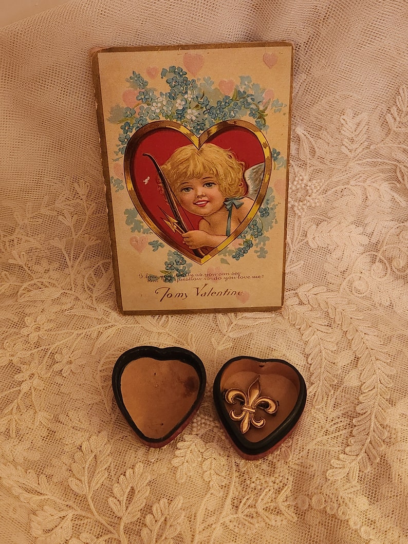 May include: A vintage Valentine's Day card with a cherub holding a bow and arrow, surrounded by flowers. The card is in a heart-shaped box with a gold fleur-de-lis charm inside.