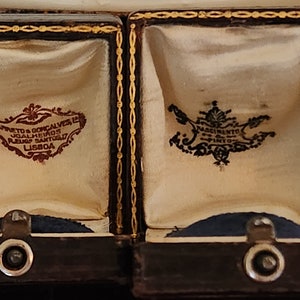 Puede incluir: Dos peque&ntilde;as cajas de madera marr&oacute;n oscuro con ribetes dorados y forro de terciopelo. Las cajas est&aacute;n abiertas y cada una tiene una etiqueta diferente en su interior. La etiqueta de la caja de la izquierda dice "Barreto & Goncalves, Joalheiros, Ruao Santo, Lisboa". La etiqueta de la caja de la derecha dice "Nascimento, Ourives, Lisboa".