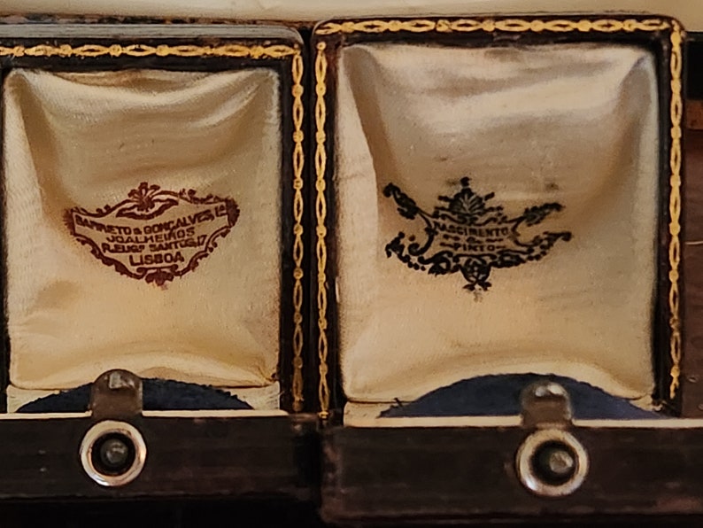 Puede incluir: Dos peque&ntilde;as cajas de madera marr&oacute;n oscuro con ribetes dorados y forro de terciopelo. Las cajas est&aacute;n abiertas y cada una tiene una etiqueta diferente en su interior. La etiqueta de la caja de la izquierda dice "Barreto & Goncalves, Joalheiros, Ruao Santo, Lisboa". La etiqueta de la caja de la derecha dice "Nascimento, Ourives, Lisboa".