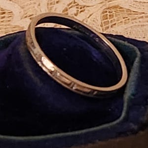 May include: A silver wedding band with a decorative pattern. The band is sitting in a blue velvet ring box.