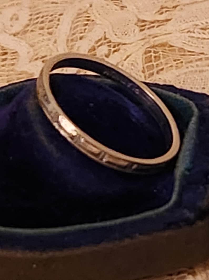 May include: A silver wedding band with a decorative pattern. The band is sitting in a blue velvet ring box.