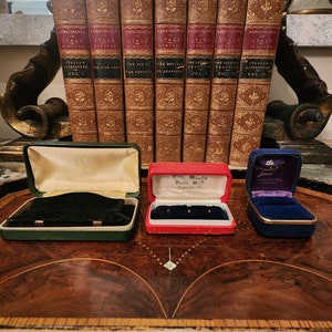 May include: A set of three vintage ring boxes. The boxes are made of green, red, and blue velvet and have gold trim. The boxes are sitting on a wooden surface. The text on the red box reads "Carl. Windel. Berlin W. 5.  Eigenthum. No. 202."