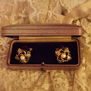 May include: A pair of gold tone earrings with a pearl in the centre of each earring. The earrings are in a purple velvet box.