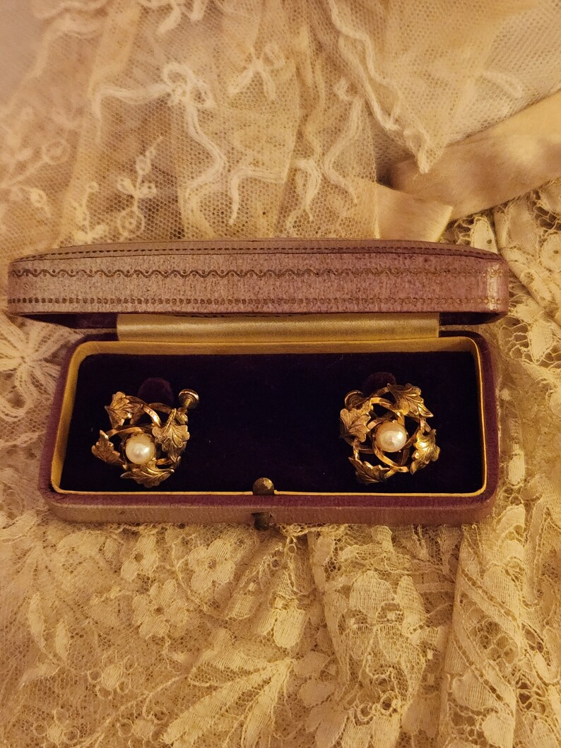 May include: A pair of gold tone earrings with a pearl in the centre of each earring. The earrings are in a purple velvet box.