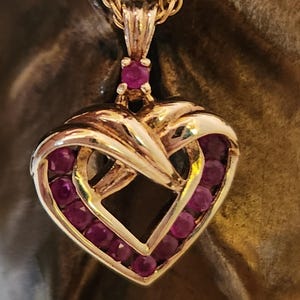 May include: A gold heart-shaped pendant necklace with a delicate chain. The pendant is adorned with a border of small, red gemstones.