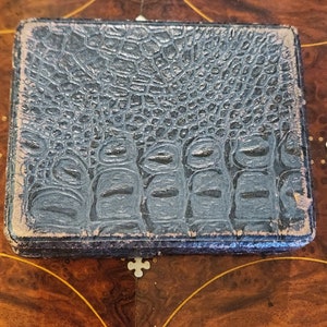 May include: A dark blue, textured, rectangular box with a crocodile skin pattern. The box is closed and has a slight sheen.