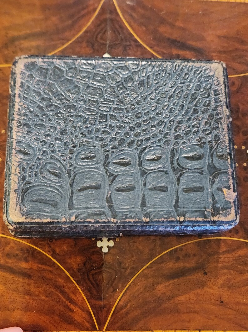May include: A dark blue, textured, rectangular box with a crocodile skin pattern. The box is closed and has a slight sheen.