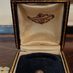 Puede incluir: Una caja de anillo de cuero marr&oacute;n con ribetes dorados y un interior de terciopelo blanco. La caja tiene una etiqueta que dice "FERREIRA BORRALHO, LISBOA".