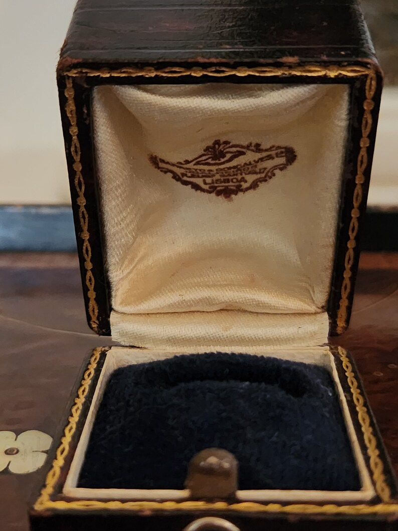 Puede incluir: Una caja de anillo de cuero marr&oacute;n con ribetes dorados y un interior de terciopelo blanco. La caja tiene una etiqueta que dice "FERREIRA BORRALHO, LISBOA".