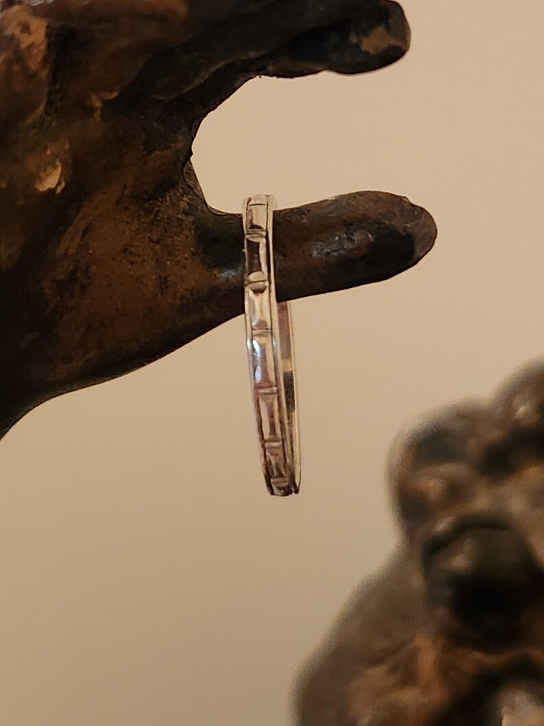 May include: A silver band ring with a textured design. The ring is thin and delicate.