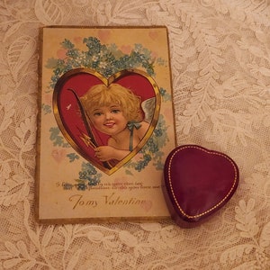 May include: Vintage Valentine's Day card with a cherub holding a bow and arrow inside a red heart. The card has a gold border and is decorated with blue forget-me-not flowers. The text on the card reads "I love you dearly as you can be But question do you love me Tomy Valentine".