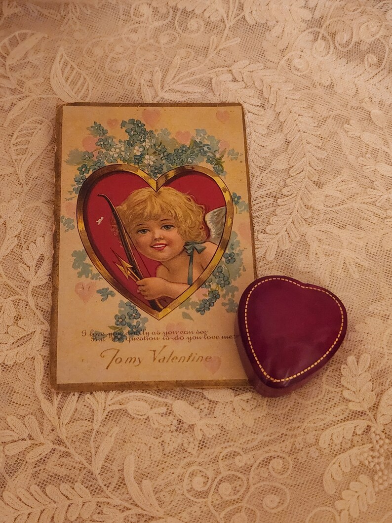 May include: Vintage Valentine's Day card with a cherub holding a bow and arrow inside a red heart. The card has a gold border and is decorated with blue forget-me-not flowers. The text on the card reads "I love you dearly as you can be But question do you love me Tomy Valentine".