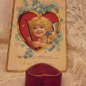 May include: A vintage Valentine's Day card with a cherub holding a bow and arrow, surrounded by blue flowers. The card has gold trim and the text "I love you dearly as you can see But the question is - do you love me? Tomy Valentine". A heart-shaped, burgundy velvet ring box sits in front of the card.