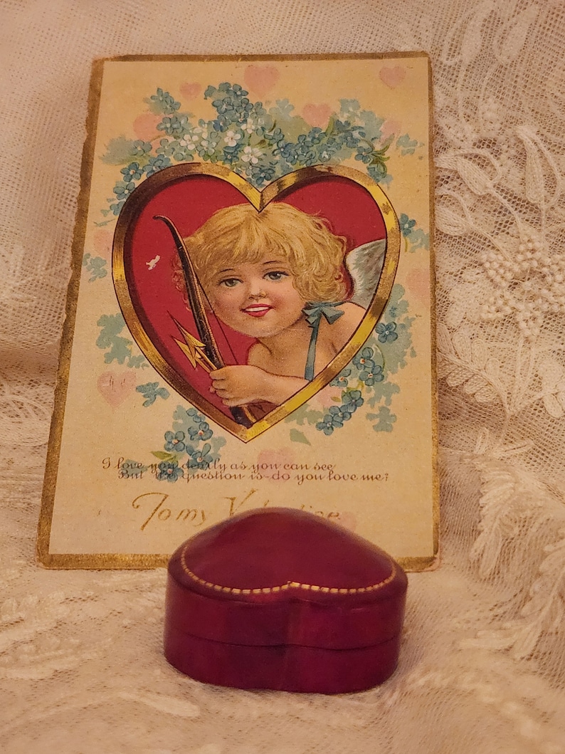 May include: A vintage Valentine's Day card with a cherub holding a bow and arrow, surrounded by blue flowers. The card has gold trim and the text "I love you dearly as you can see But the question is - do you love me? Tomy Valentine". A heart-shaped, burgundy velvet ring box sits in front of the card.