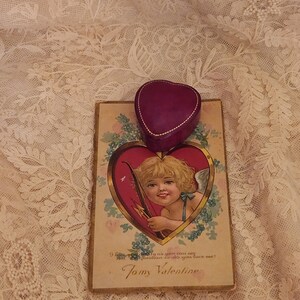 May include: A vintage Valentine's Day card with a cherub holding an arrow and a heart-shaped box with a gold trim. The card has a gold border and the text "9 love modely as you eau deg estion to do you love me? Tomy Valentine".