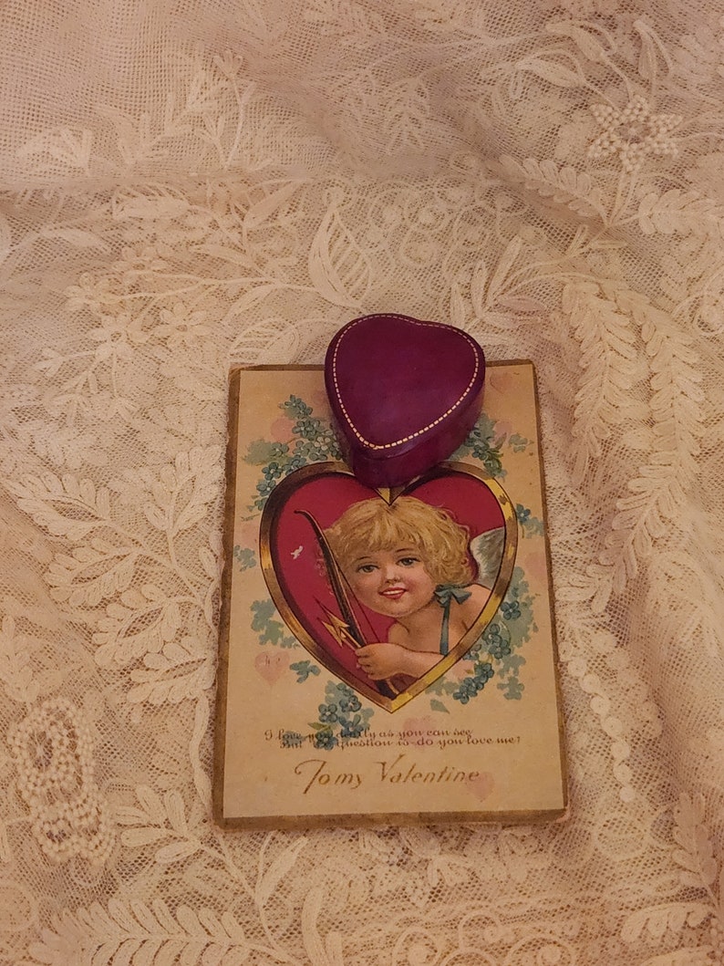 May include: A vintage Valentine's Day card with a cherub holding an arrow and a heart-shaped box with a gold trim. The card has a gold border and the text "9 love modely as you eau deg estion to do you love me? Tomy Valentine".