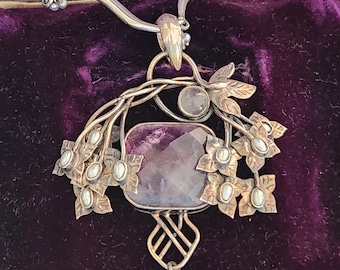 May include: A silver necklace with a large, rectangular, purple gemstone pendant. The pendant is adorned with floral and leaf accents, small pearls, and a large, pear-shaped pearl drop. The necklace is displayed on a purple velvet surface.