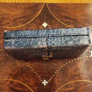 May include: A black leather-covered box with a hinged lid and a gold metal clasp. The box is closed and sitting on a wooden surface.