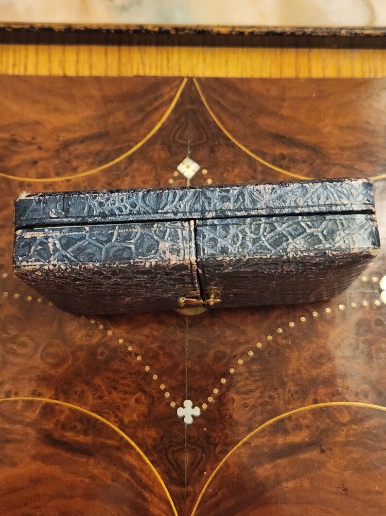 May include: A black leather-covered box with a hinged lid and a gold metal clasp. The box is closed and sitting on a wooden surface.