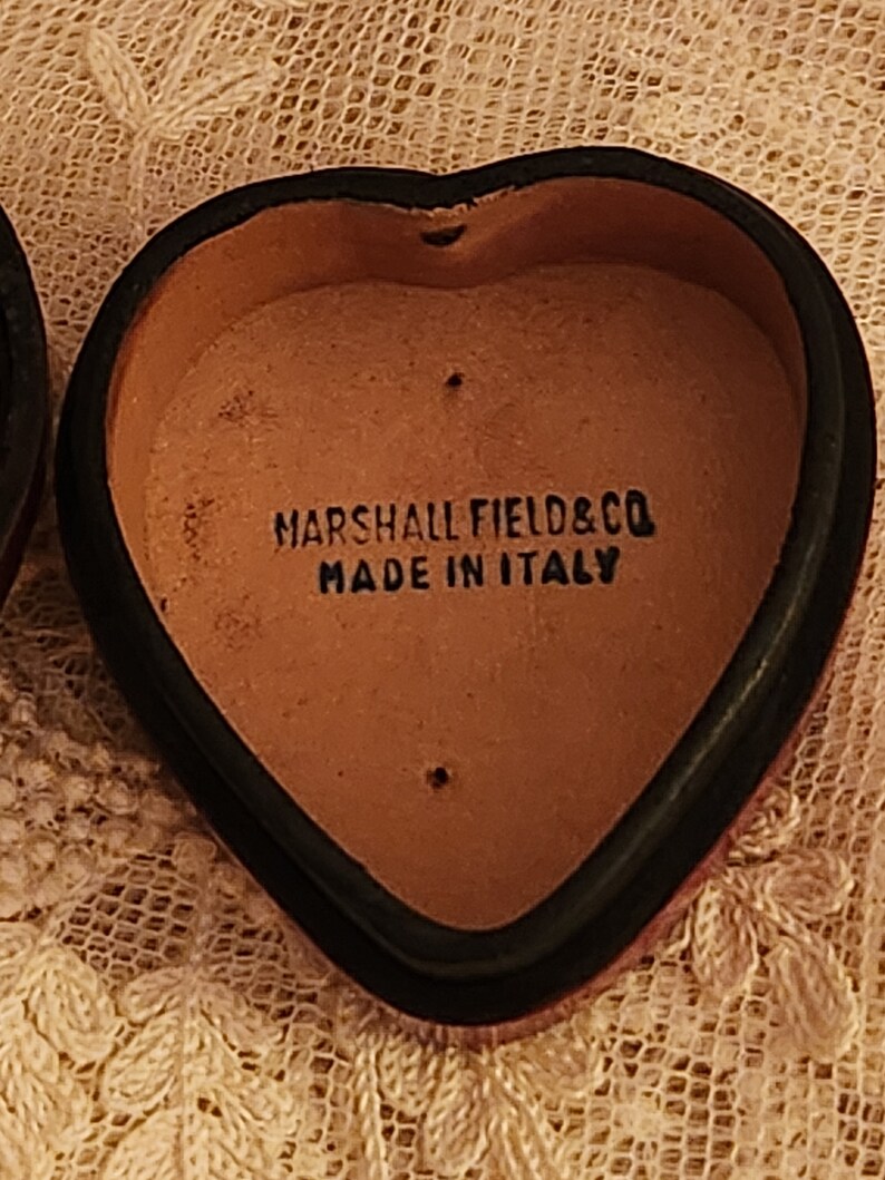 May include: A heart-shaped leather box with a black rim. The inside of the box is brown and has the text "MARSHALL FIELD & CO MADE IN ITALY" printed on it.