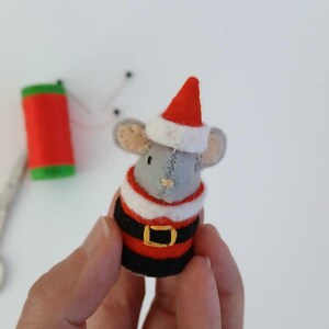 May include: A handmade felt mouse wearing a Santa Claus outfit, complete with a red hat and black belt with a gold buckle. The mouse is gray with tan ears. A red spool of thread, scissors, and pins are in the background.