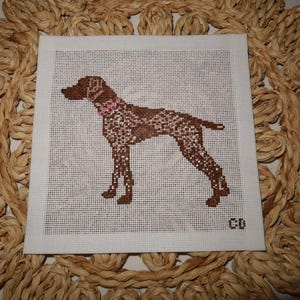May include: A square white fabric piece featuring a cross-stitch embroidery of a brown and white spotted dog in profile. The dog wears a pink collar, and the initials CD are stitched in the bottom right corner on the white grid background.