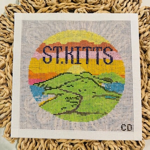 May include: A needlepoint canvas featuring the text "ST. KITTS" in navy blue, set against a colorful sunset design. The image includes a green island silhouette and is framed by a woven, circular mat.