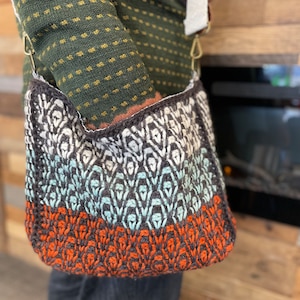 Puede incluir: Bolso de hombro hecho a mano con un patrón geométrico en tonos blanco, gris, turquesa y naranja. El bolso tiene una correa color crema y herrajes color latón. El bolso está hecho de material de punto.