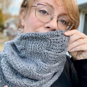 Può includere: Un collo a lupetto di maglia grigia con un motivo a treccia. Il collo a lupetto è indossato da una persona con occhiali e capelli biondi.