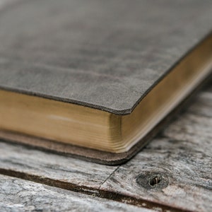 Puede incluir: Primer plano de un diario encuadernado en cuero gris con un borde dorado. El diario est&aacute; apoyado sobre una superficie de madera desgastada.
