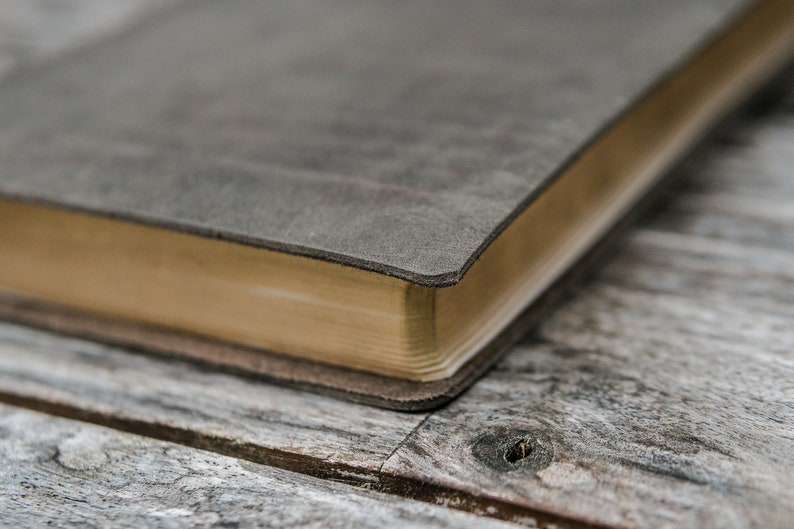 Puede incluir: Primer plano de un diario encuadernado en cuero gris con un borde dorado. El diario est&aacute; apoyado sobre una superficie de madera desgastada.