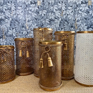 May include: Six gold metal decorative containers with a mesh design and tassel accents. The containers are arranged in a row on a brown surface.