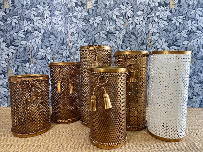 May include: Six gold metal decorative containers with a mesh design and tassel accents. The containers are arranged in a row on a brown surface.