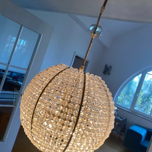 May include: A large, round, crystal chandelier with gold accents. The chandelier is made up of many small, clear crystals that are arranged in a geometric pattern. The chandelier is hanging from a ceiling and is illuminated by a light bulb.